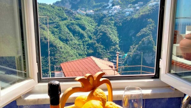 A table with a bottle of wine, a bowl of lemons, and a glass of water, with a scenic view of a mountainous landscape through a window. - Independent house with access to a panoramic lemon grove on the Amalfi Coast! (Scala)