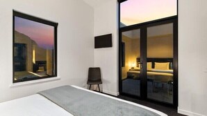 A modern hotel room with a large bed, a chair, a TV, and a sliding glass door leading to a balcony with a view of the sunset. - Penthouse Leisure at La Residence du Parc (Queenstown)