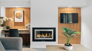 A modern living room with a fireplace, a flat-screen TV mounted on the wall, a desk with a chair, and a potted plant. - Penthouse Leisure at La Residence du Parc (Queenstown)