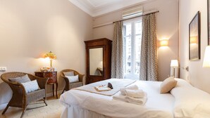 Een hotelkamer met een groot bed, een rieten stoel, een houten dressoir en een raam met gordijnen.