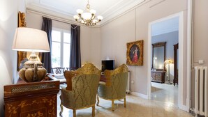 Een kamer met een kroonluchter, een tafel met een lamp, twee sierlijke stoelen, een lijstwerk schilderij en een spiegel.