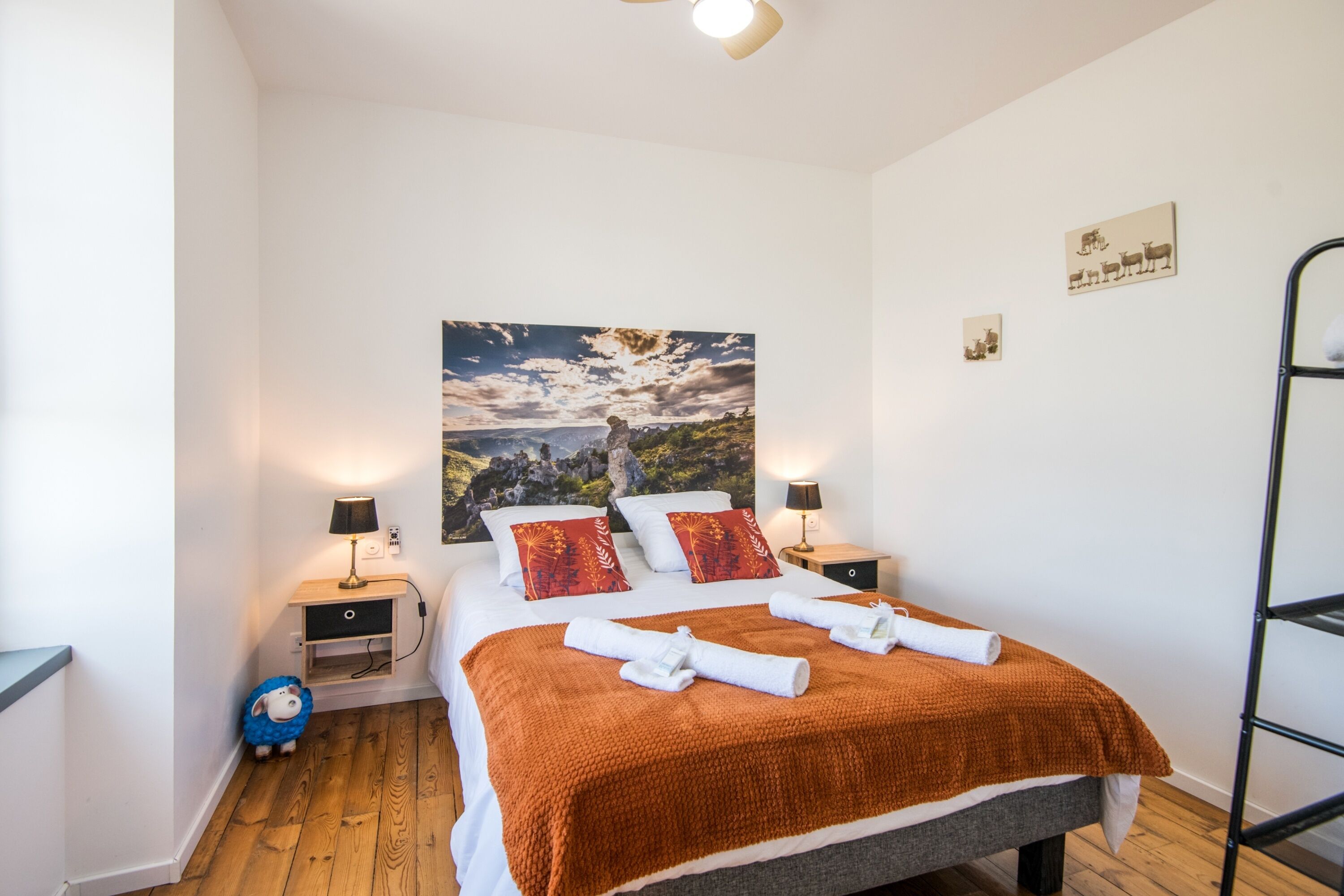 A neatly made bed with a brown blanket, two bedside lamps, a small nightstand, a wall-mounted shelf, and a framed picture of a landscape.