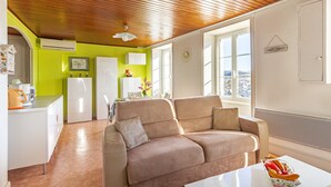 A modern living space with a wooden ceiling, a beige sofa, a white coffee table, and a kitchen area with a refrigerator and a sink. - Le Barry - Studio avec jardin (Murat)