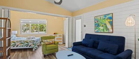 A room with a bed, a blue sofa, a green armchair, a small table, a ceiling fan, and a window with a view of greenery.