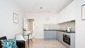 Een moderne keuken met grijze kastjes, een zwart aanrechtblad en een ingebouwde oven. Er is een eethoek met een glazen tafel en stoelen, en een zwarte bank met een kleurrijk kussen.