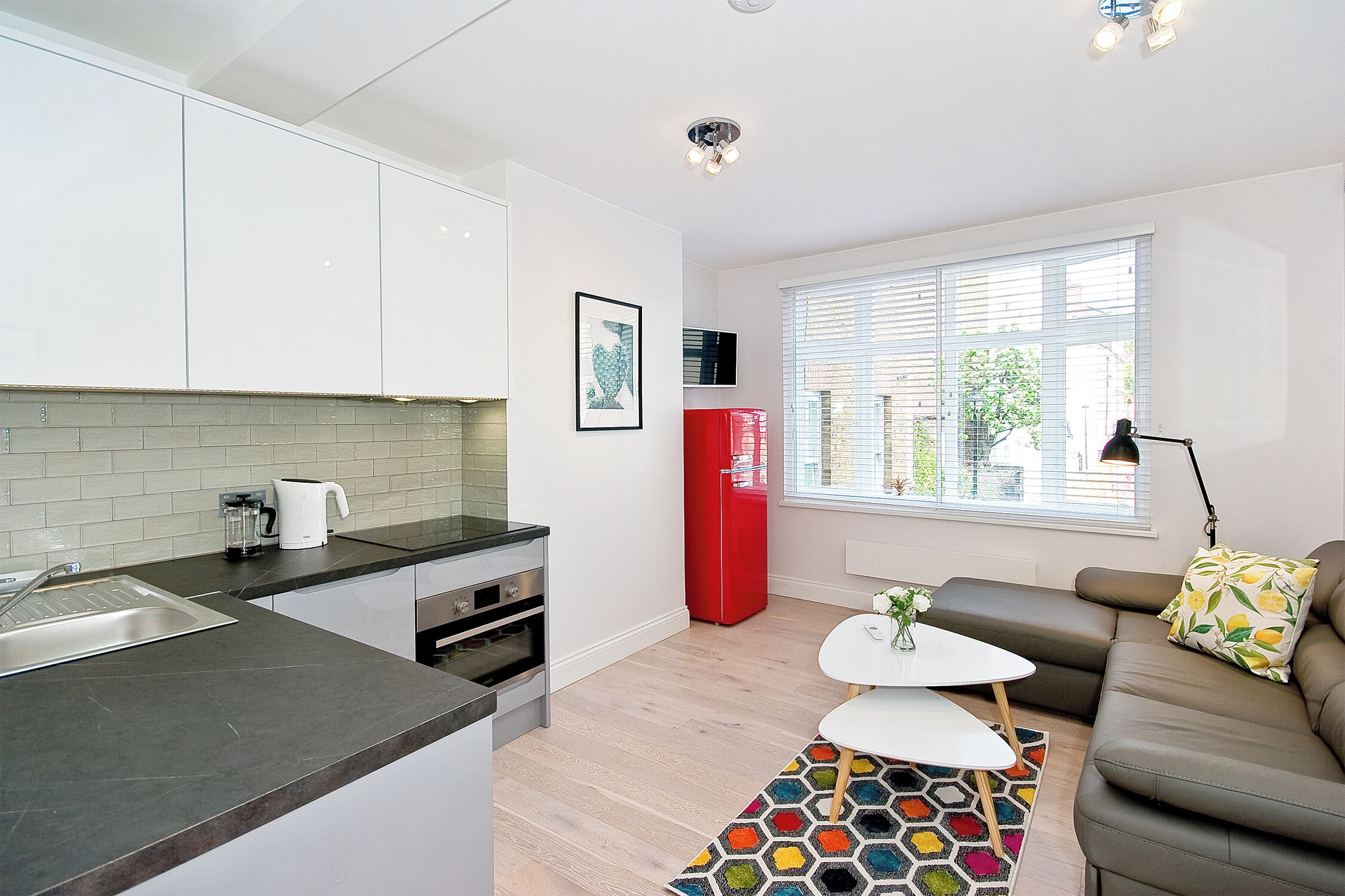 Eine moderne Küche mit Spüle, Herd und einem roten Kühlschrank. Ein weißer Esstisch, ein farbenfroher Teppich und eine braune Couch mit Kissen sind ebenfalls vorhanden.