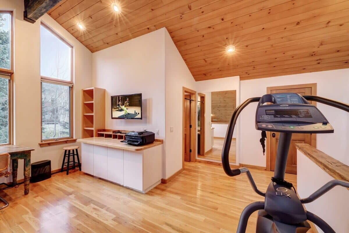 A spacious room with a wooden ceiling, a treadmill, a TV mounted on the wall, and a kitchen area with a counter and stools.