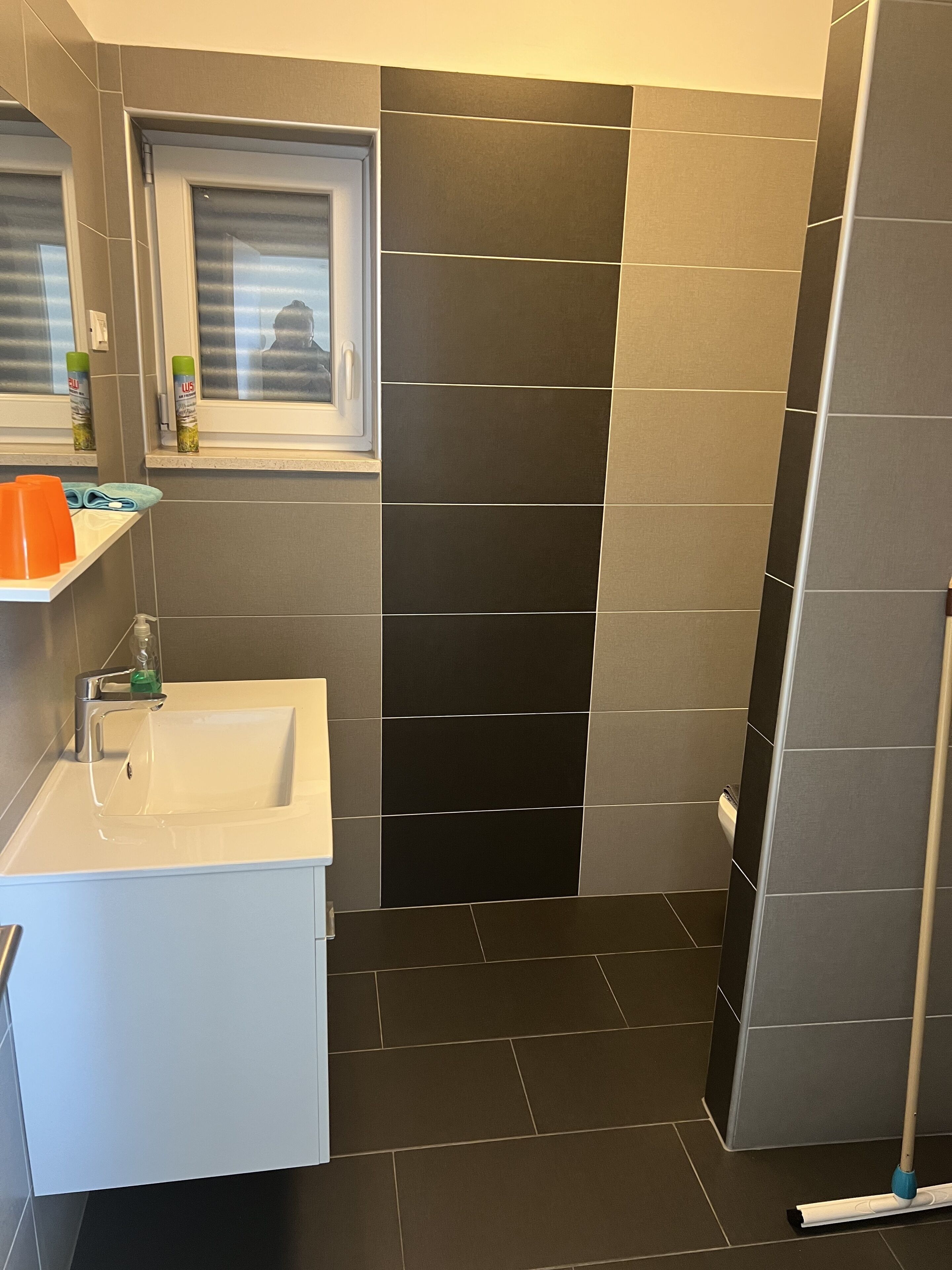 A modern bathroom with a white sink, a window above it, and a shower area with dark tiles.