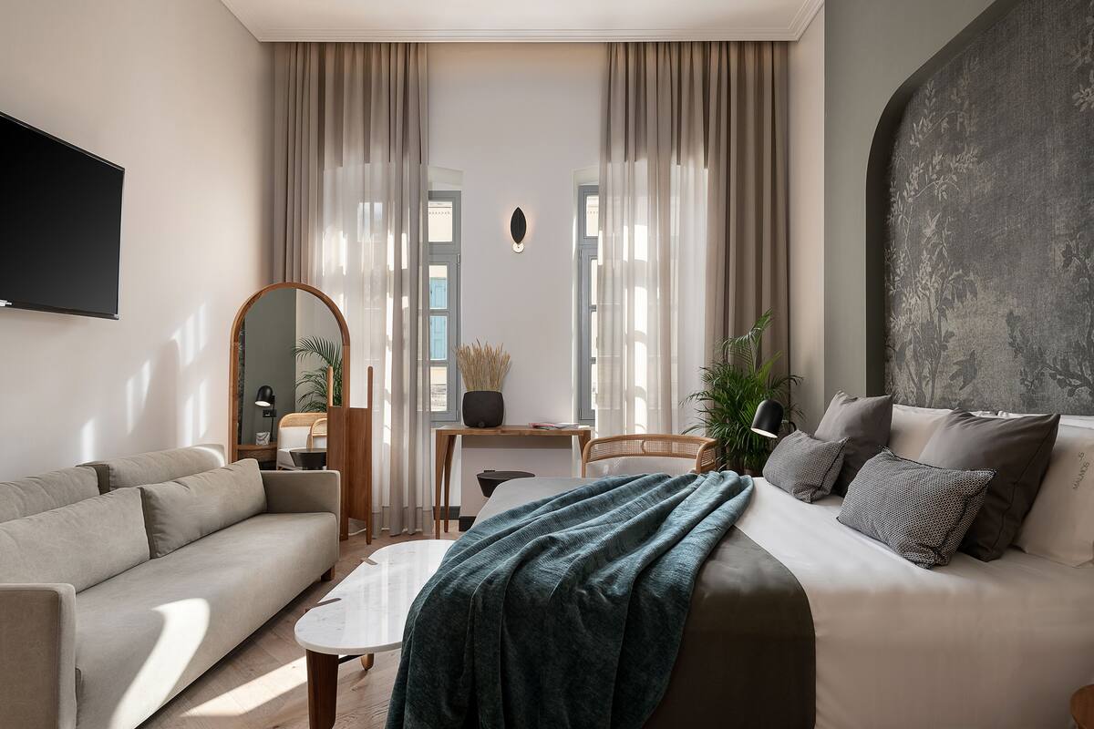 A modern hotel room with a sofa, a bed with a blanket, a desk with a lamp, and a large mirror.