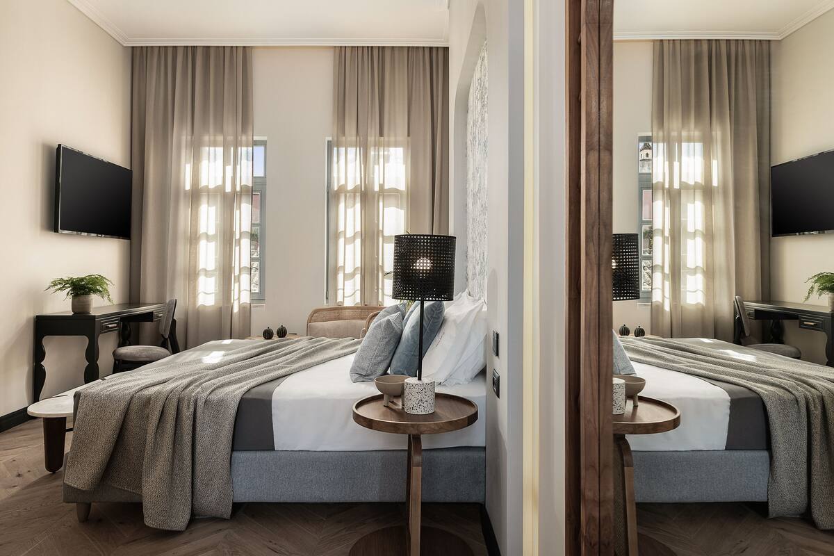 A modern hotel room with a large bed, a small table with a lamp, and a TV mounted on the wall.