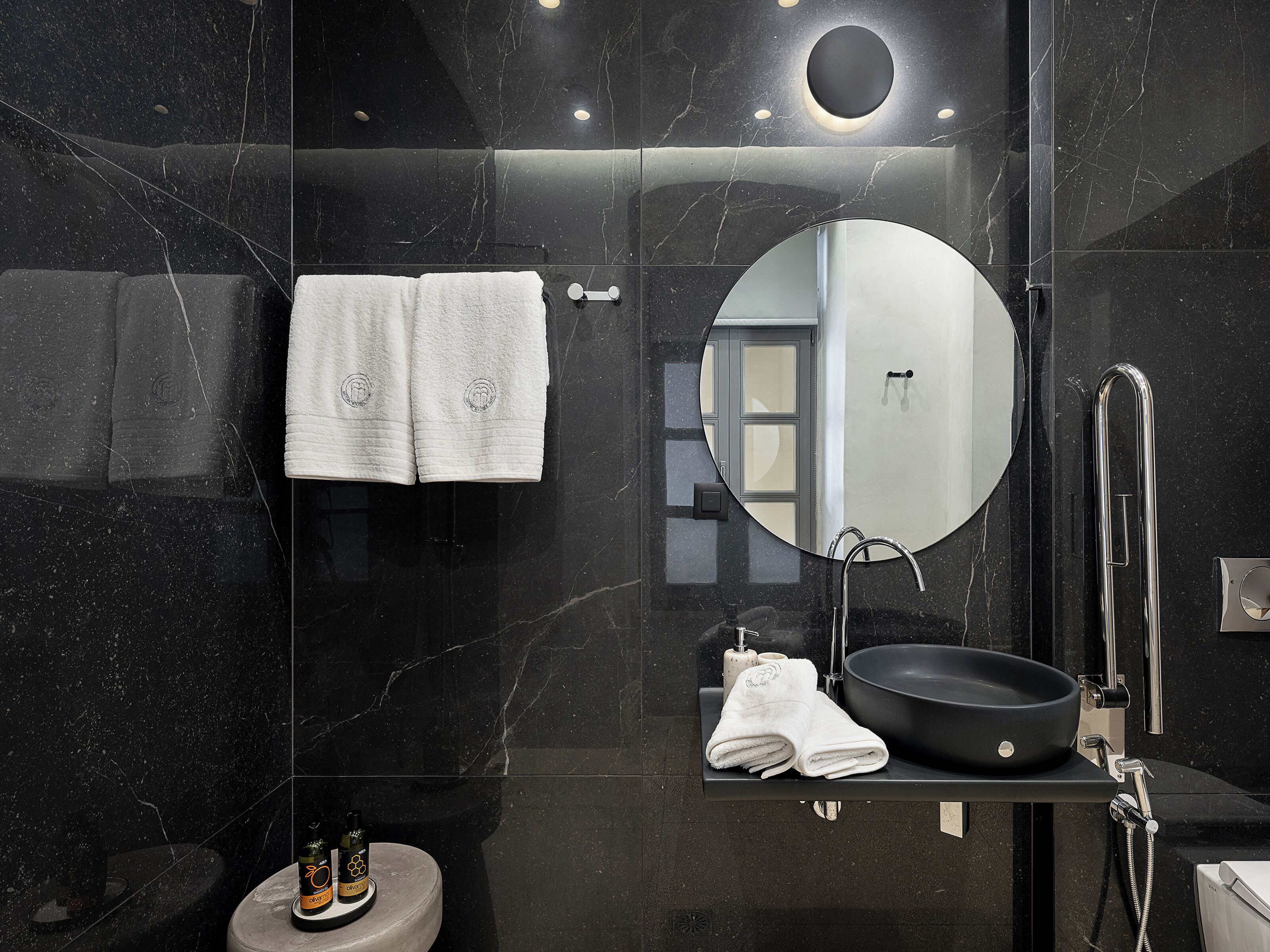 Un baño moderno con pared de mármol negro, un espejo redondo, un lavabo negro y dos toallas blancas.