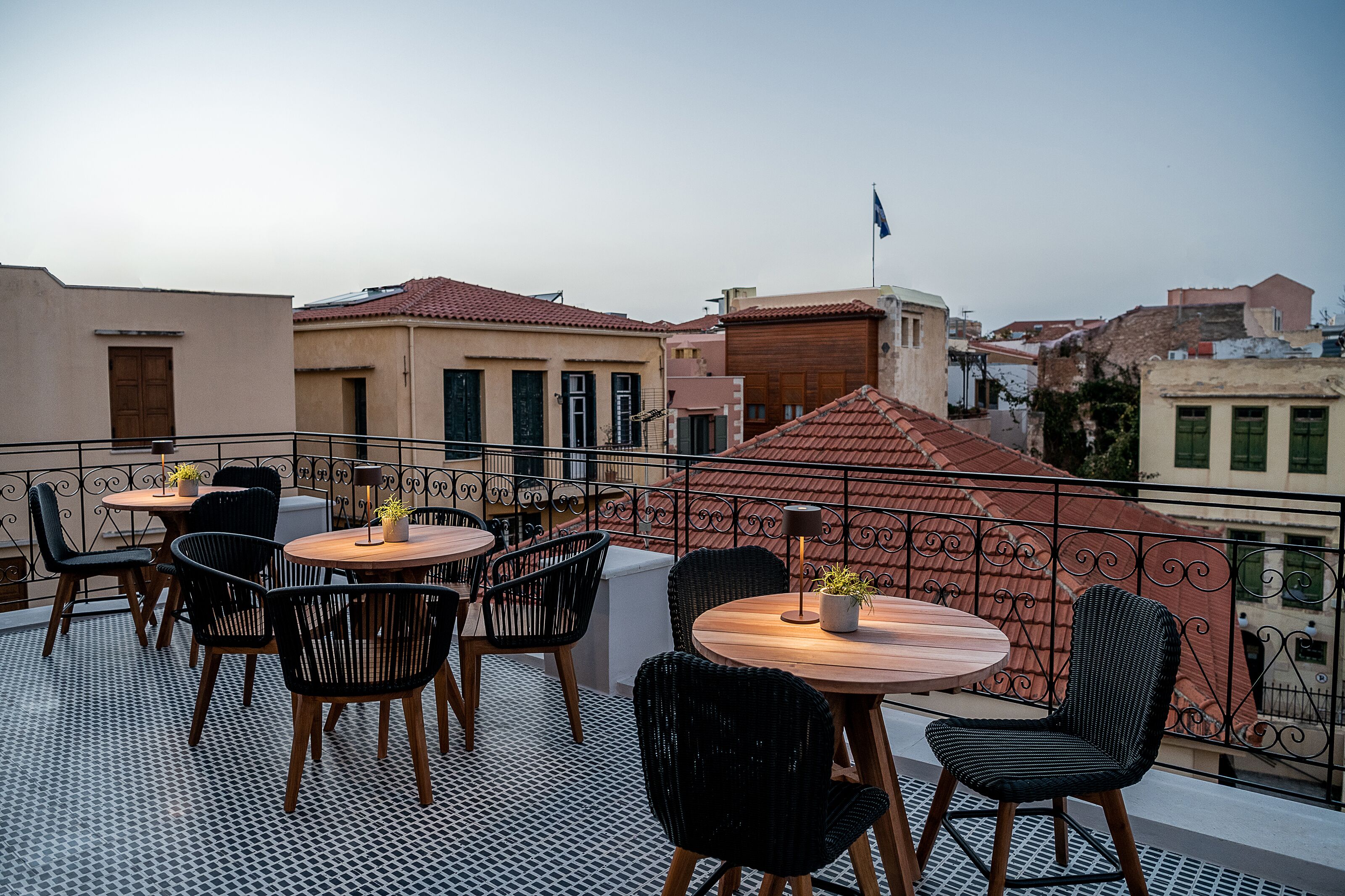 A rooftop terrace with round tables and chairs, a patterned floor, and a view of surrounding buildings.