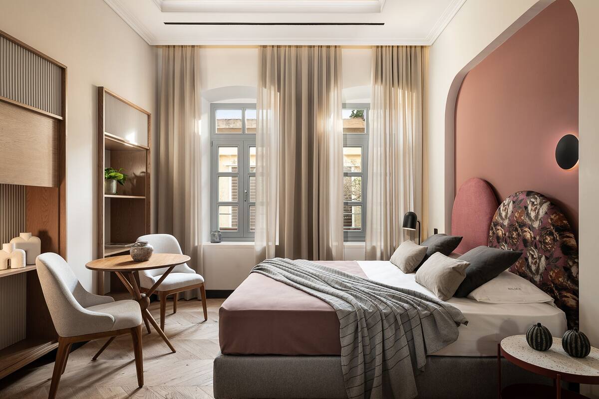A modern bedroom with a bed, a desk, and chairs. There are curtains on the windows, a plant on the desk, and a lamp on the bedside table.