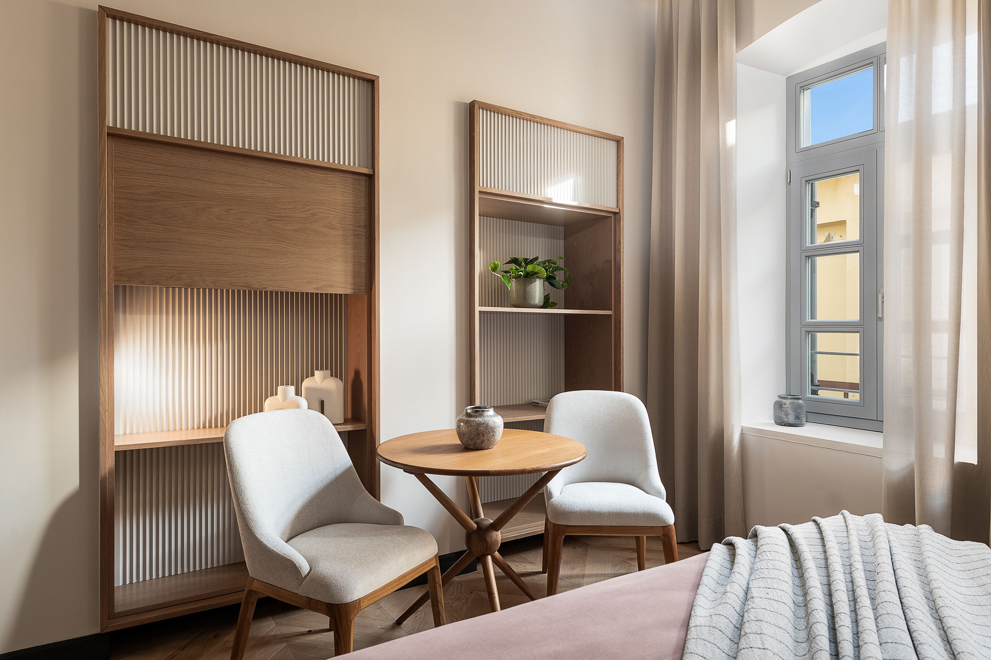Una habitación moderna con una cama, dos sillas, una mesa redonda de madera y estanterías empotradas con una planta.