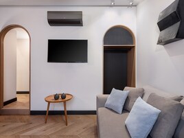 Un salón moderno con una televisión de pantalla plana montada en la pared, una mesa de centro de madera con objetos decorativos, un sofá color beige con cojines y un equipo de aire acondicionado empotrado.