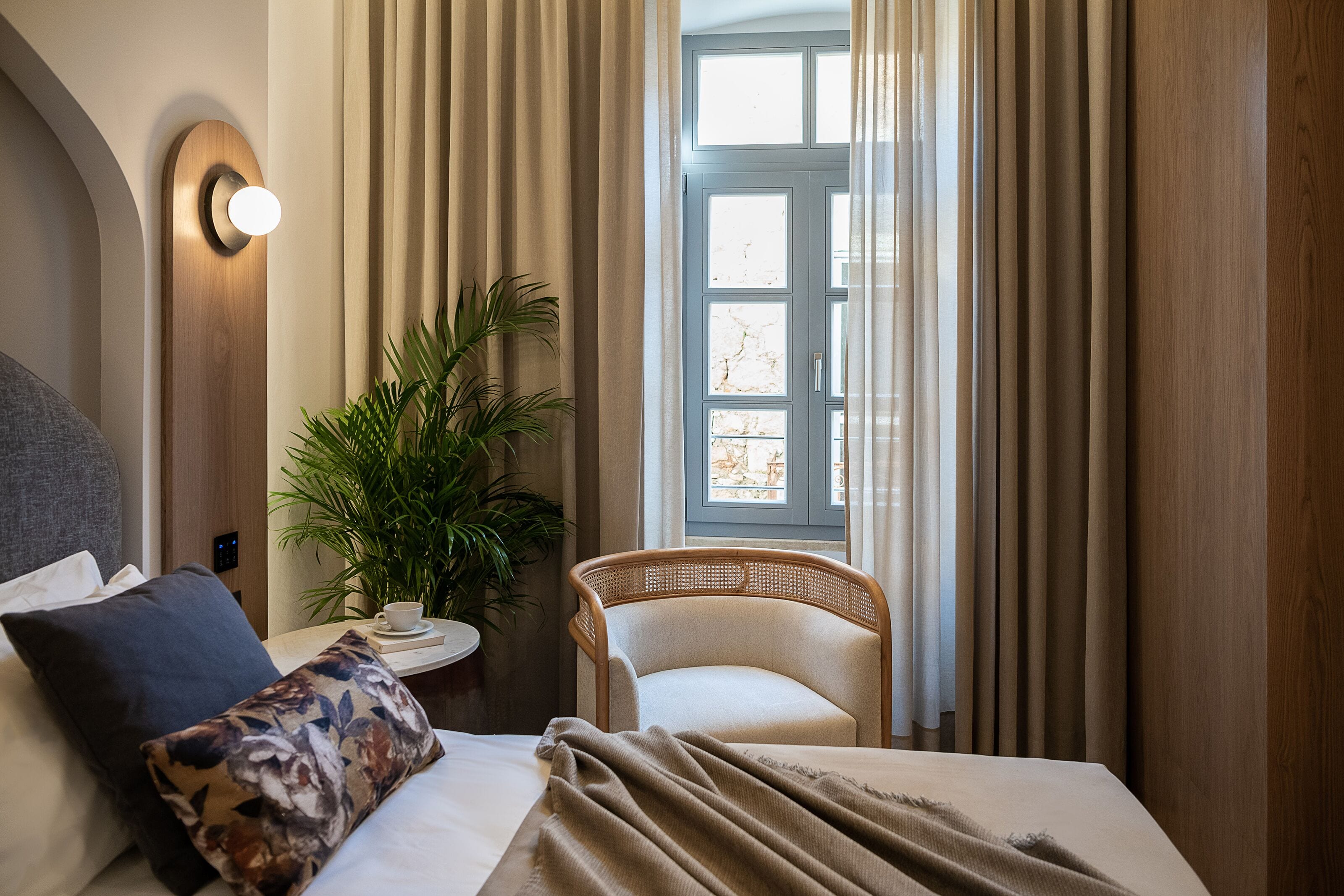 A hotel room with a bed, a chair, a potted plant, and a window with curtains.