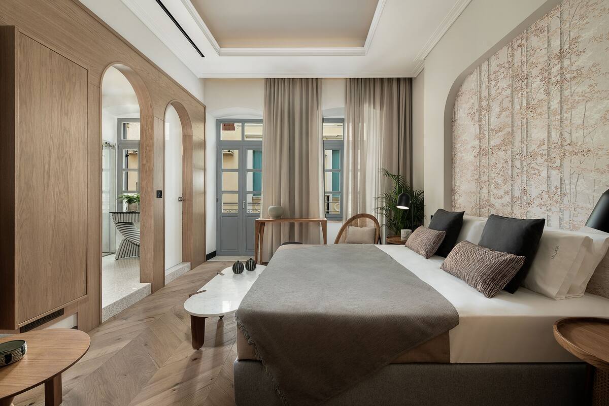 A modern hotel room with a large bed, a sitting area with a sofa and armchairs, a small table, and a view of the outside balcony.