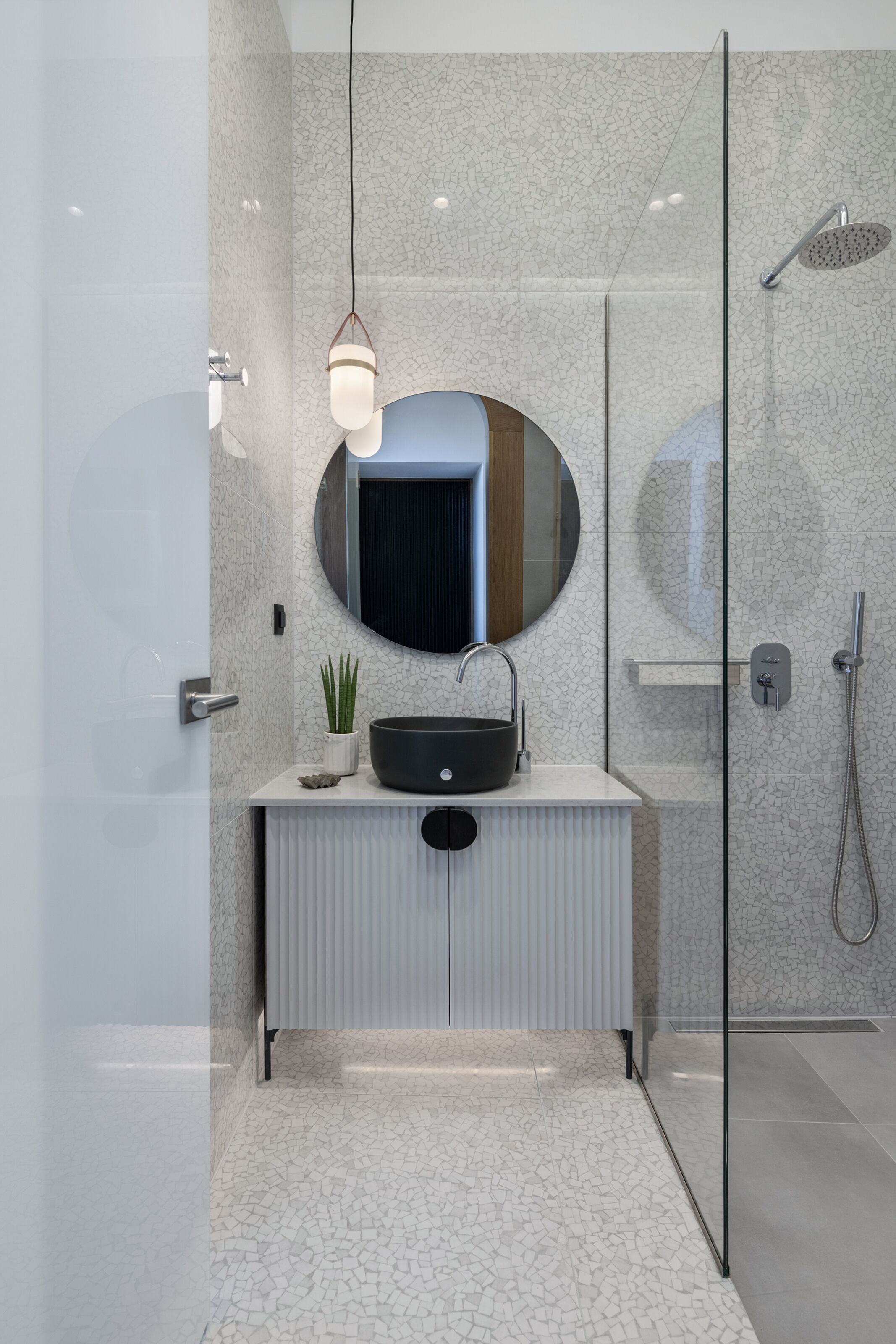 A modern bathroom with a glass shower enclosure, a round mirror, a black sink, and a pendant light.