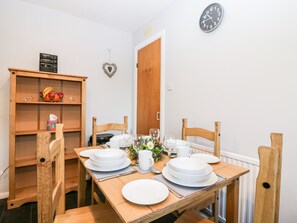 A dining area with a wooden table set for four, surrounded by wooden chairs. A wooden cabinet with a fruit bowl and glasses is to the left. A clock is on the wall above the door.