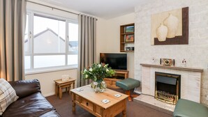 A living room with a wooden coffee table, a leather sofa, a television, a fireplace, and a large window with curtains. - 10 Annesley Park (Banchory)