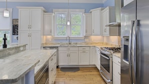 A modern kitchen with white cabinets, a stainless steel refrigerator, a built-in oven, and a large window providing natural light. - Aqua Waters - Newly Updated Vacation Home with Golf Cart, Private Pool + Cabana! (Destin)