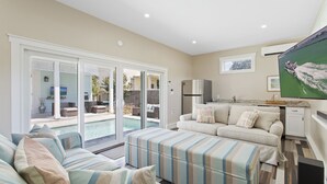 A spacious living room with a large sofa, a striped ottoman, and a television mounted on the wall. - Aqua Waters - Newly Updated Vacation Home with Golf Cart, Private Pool + Cabana! (Destin)