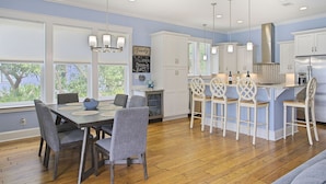 A modern kitchen with white cabinets, a central island with bar stools, and a dining area with a table and chairs. - Aqua Waters - Newly Updated Vacation Home with Golf Cart, Private Pool + Cabana! (Destin)