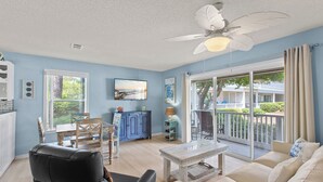 Un salon spacieux doté d’une grande fenêtre, d’un ventilateur de plafond, d’un téléviseur et d’un balcon aménagé avec du mobilier d’extérieur.