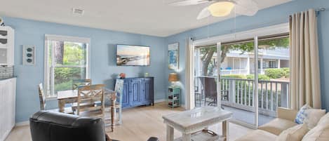 A spacious living room with a large window, a ceiling fan, a television, and a balcony with outdoor furniture.