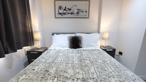 Ein ordentlich bezogenes Bett mit einer flauschigen, weißen und grauen Decke, zwei Nachttischlampen und einem gerahmten Kunstwerk an der Wand.