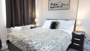 Ein ordentlich bezogenes Bett mit einer flauschigen, weißen und grauen Decke, zwei Nachttischlampen, einem schwarzen Nachttisch und einem gerahmten Kunstwerk an der Wand.