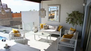 Un espacio al aire libre iluminado por el sol con un sofá blanco, una mesa de vidrio y una lámpara colgante.
