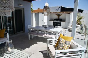 Un espace extérieur aménagé en terrasse, avec une table à manger, des chaises blanches et un canapé avec des coussins.