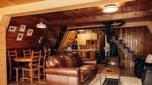 Un acogedor espacio tipo loft con una zona de estar que incluye un sofá de cuero marrón, una mesa de comedor de madera y sillas. Se ve una pequeña cocina de madera con estufa al fondo.