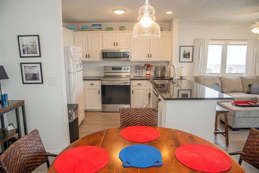 Une cuisine moderne avec des meubles blancs, un four à micro-ondes, une cuisinière et un coin repas comprenant une table en bois et des sets de table rouges.