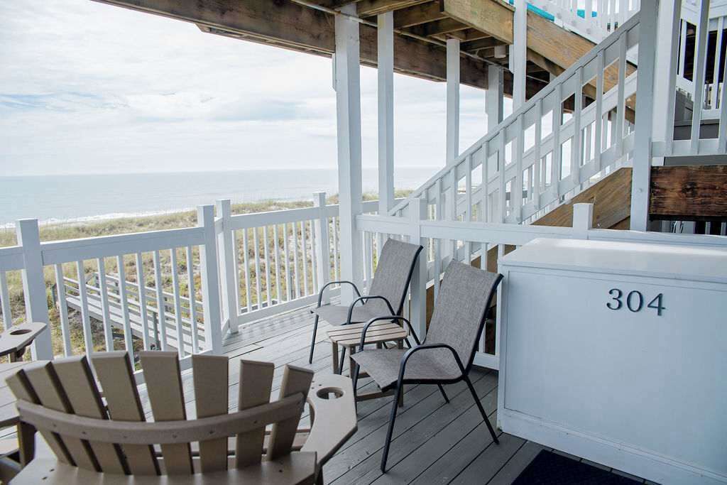 A covered outdoor seating area with a view of the beach, wooden chairs, a table, and a white cooler marked with the number 304.