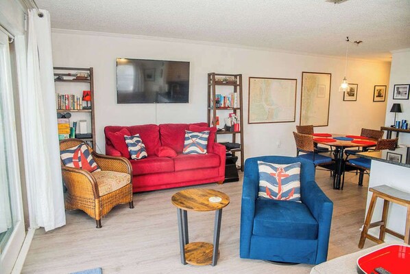 Una sala de estar con un sofá rojo, un sillón azul, sillas de mimbre, una pequeña mesa de madera y un comedor con sillas rojas y azules.