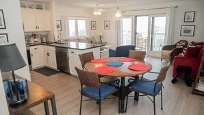 Una cocina moderna con gabinetes blancos, una zona de comedor con una mesa redonda y sillas azules, y un balcón con mobiliario de mimbre.