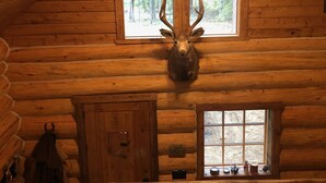 Interno di una baita di legno con una grande testa di cervo appesa al muro, una ventola a soffitto e una finestra che si affaccia sulla foresta.