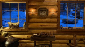 Interno accogliente di una baita con pareti in legno, un tavolo centrale con candele e finestre che si affacciano su un paesaggio innevato.