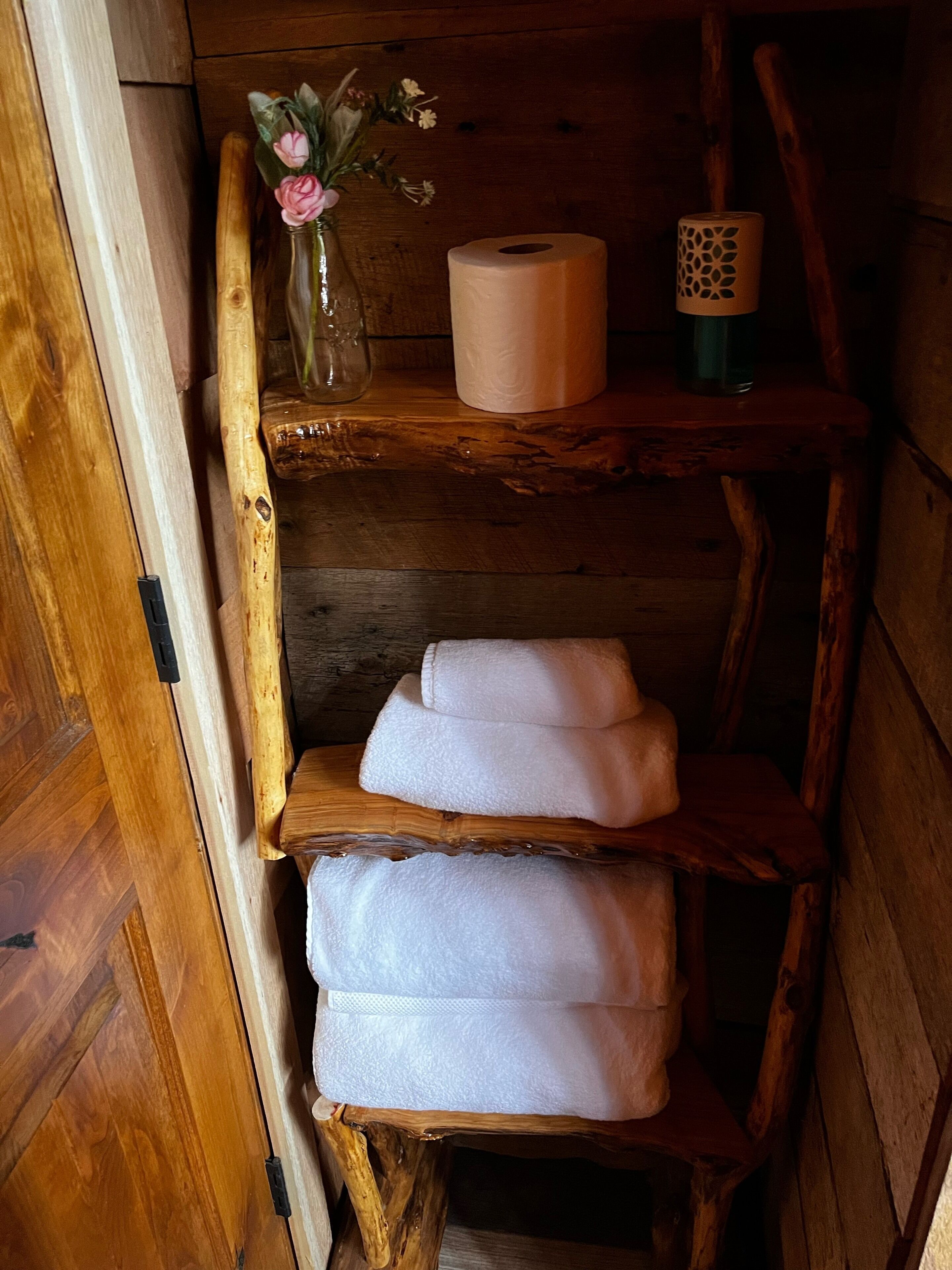 A wooden shelf with a vase of flowers, a roll of toilet paper, and folded towels.