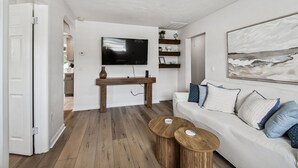 A modern living room with a wooden floor, a white sofa with blue and white cushions, a coffee table, a TV mounted on the wall, and a painting above the sofa.