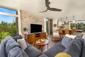 A modern living room with a large TV, grey sofas, a wooden coffee table, and a stone wall. - Breezy + Spacious 3/2 Home with Bali Hai View + Hot Tub (Princeville)