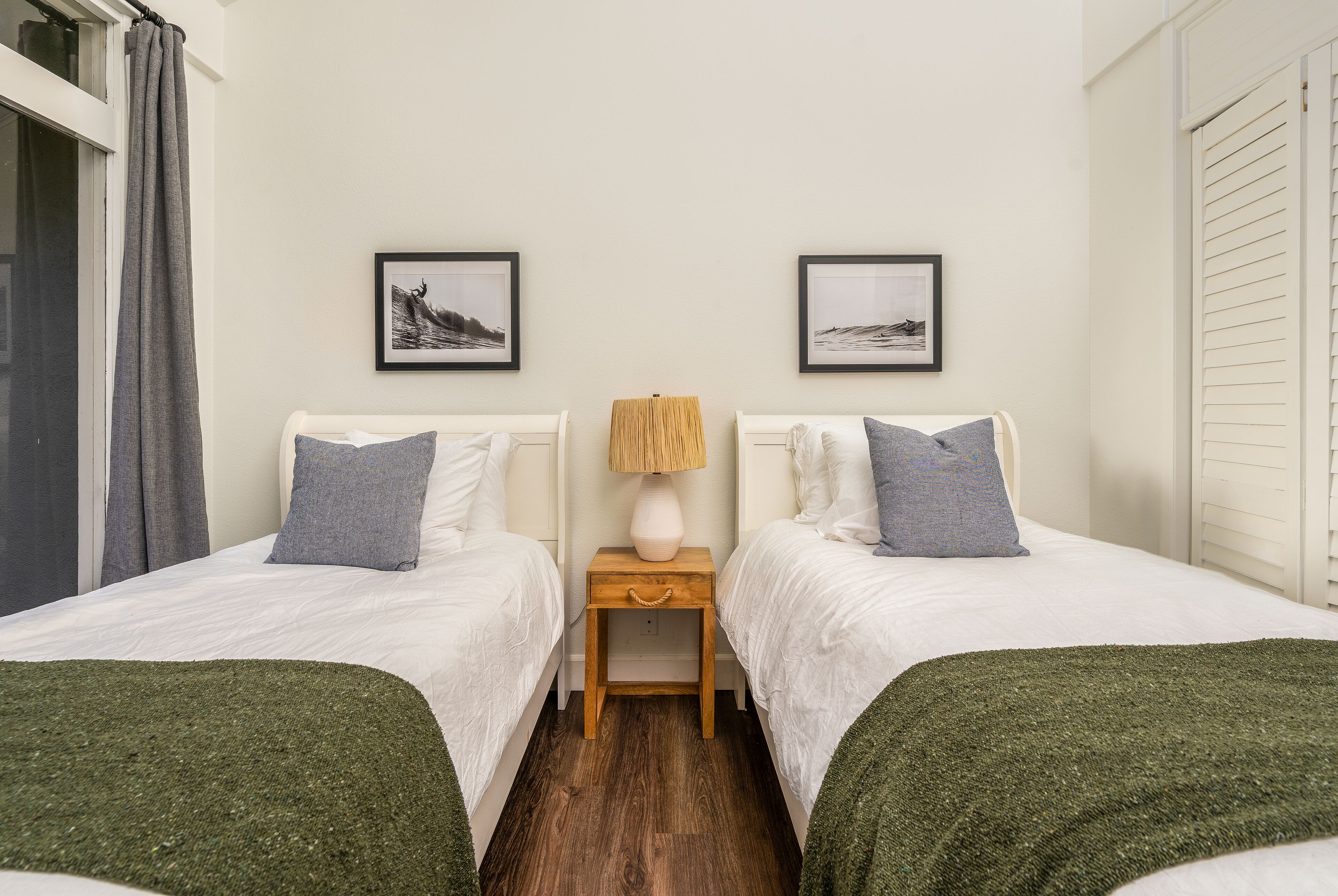 Hotellihuone, jossa on kaksi erillistä sänkyä, yöpöytä lampun kanssa ja kaksi kehystettyä kuvaa seinällä.