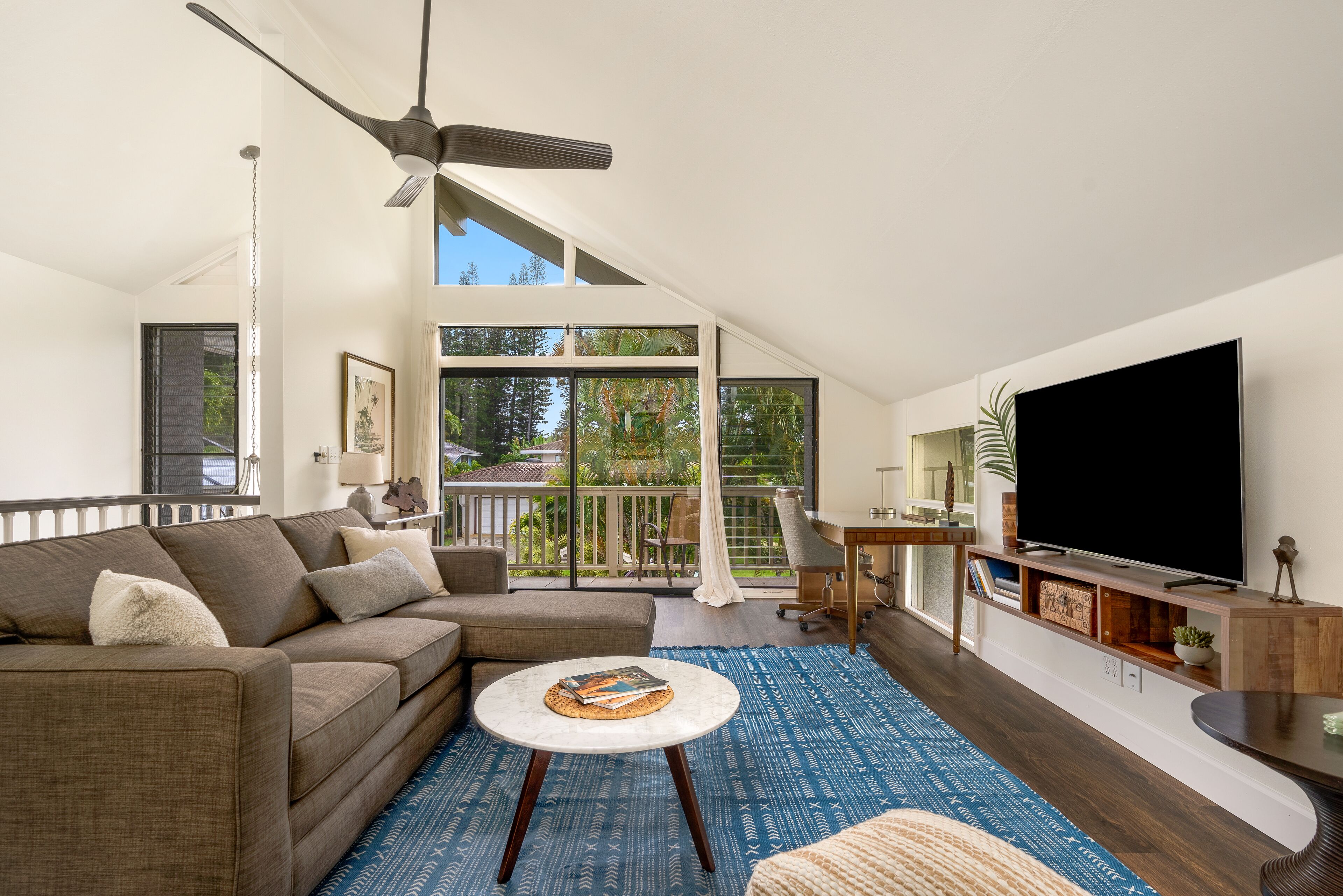 A modern living room with a large TV, a sofa, a coffee table, and a balcony with a view of trees.