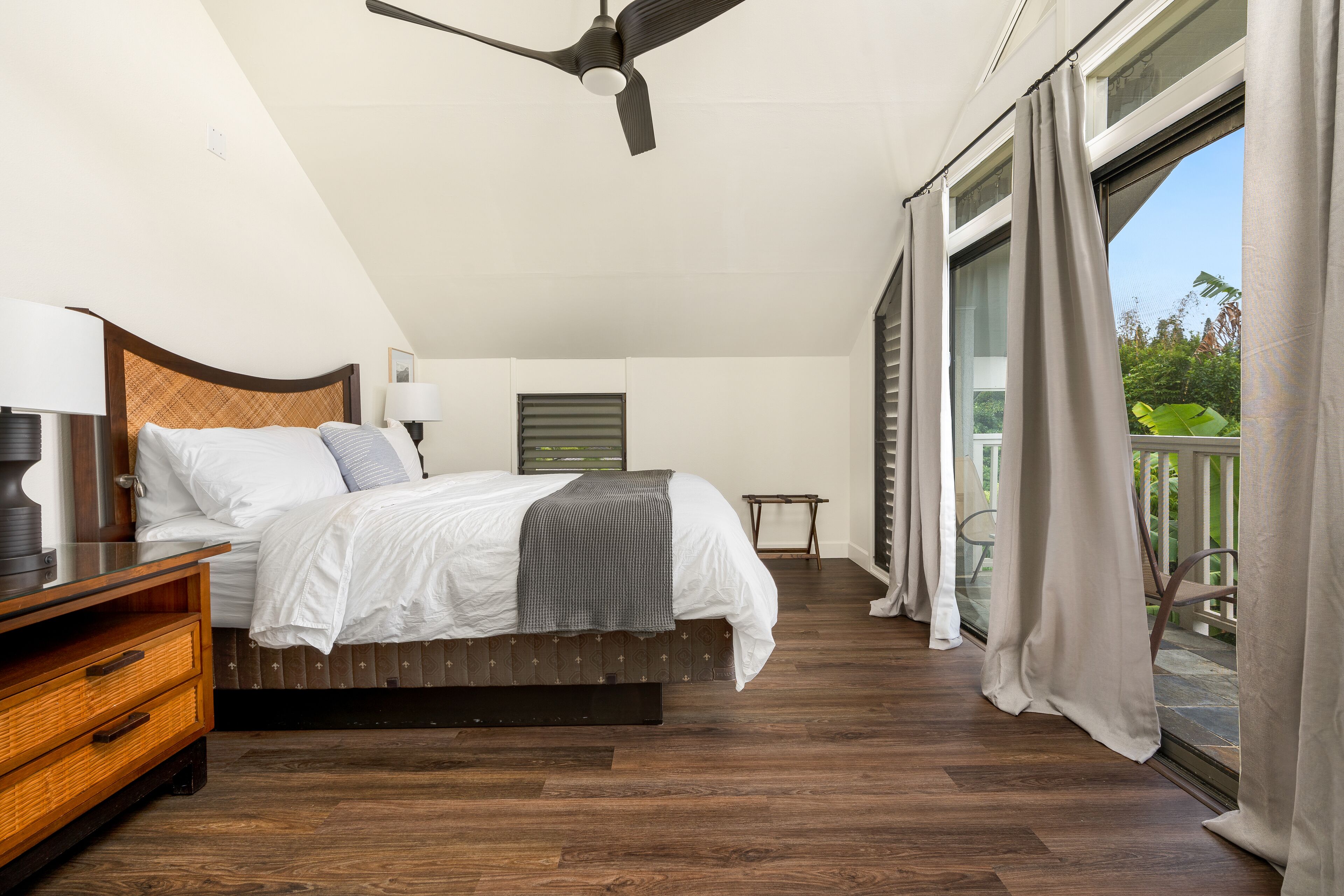 A bedroom with a bed, bedside tables, a ceiling fan, and a view through a large window.