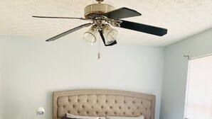 Ein Schlafzimmer mit einer gesteppten Kopfteile, einem Bett mit heller Bettdecke, einem Nachttisch mit Lampe und einem Deckenventilator.
