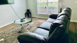 Ein Wohnzimmer mit einem schwarzen Ledersofa, einem Glastisch und einem an der Wand befestigten Flachbildfernseher.