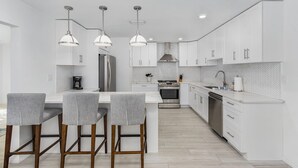 A modern kitchen with white cabinetry, a central island with bar stools, stainless steel appliances, and pendant lighting. - Newly Updated!  RiverView Retreat  Historic Fort Myers, FL 10 guests, Spa & pool (Fort Myers)