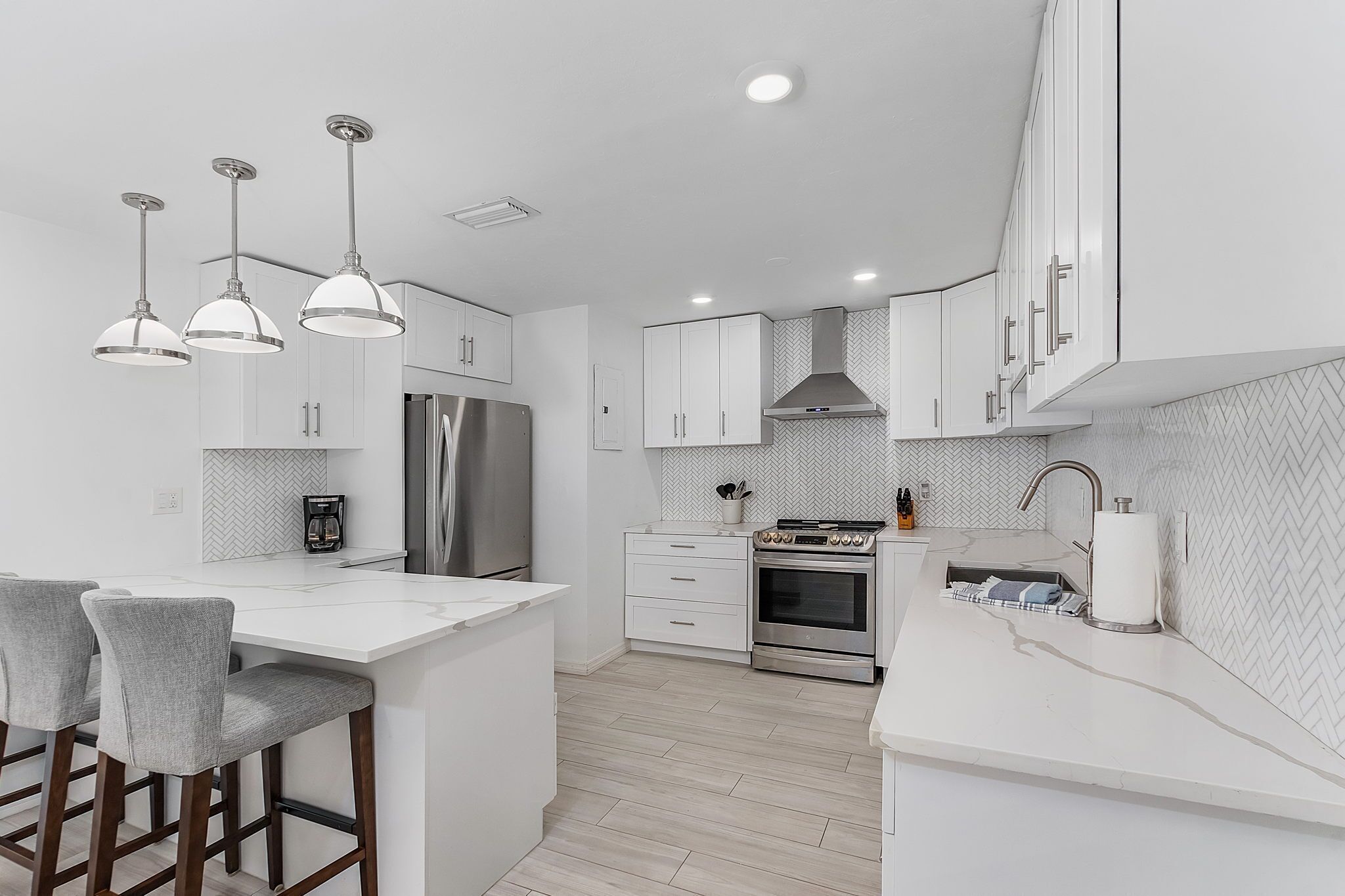 A modern kitchen with white cabinetry, a stainless steel refrigerator, an oven, and a coffee maker. There are three pendant lights and a bar area with stools.